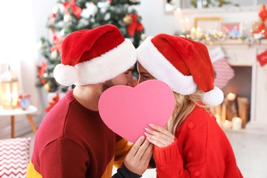 Young couple in Santa hats hiding behind heart at home. Christmas celebration