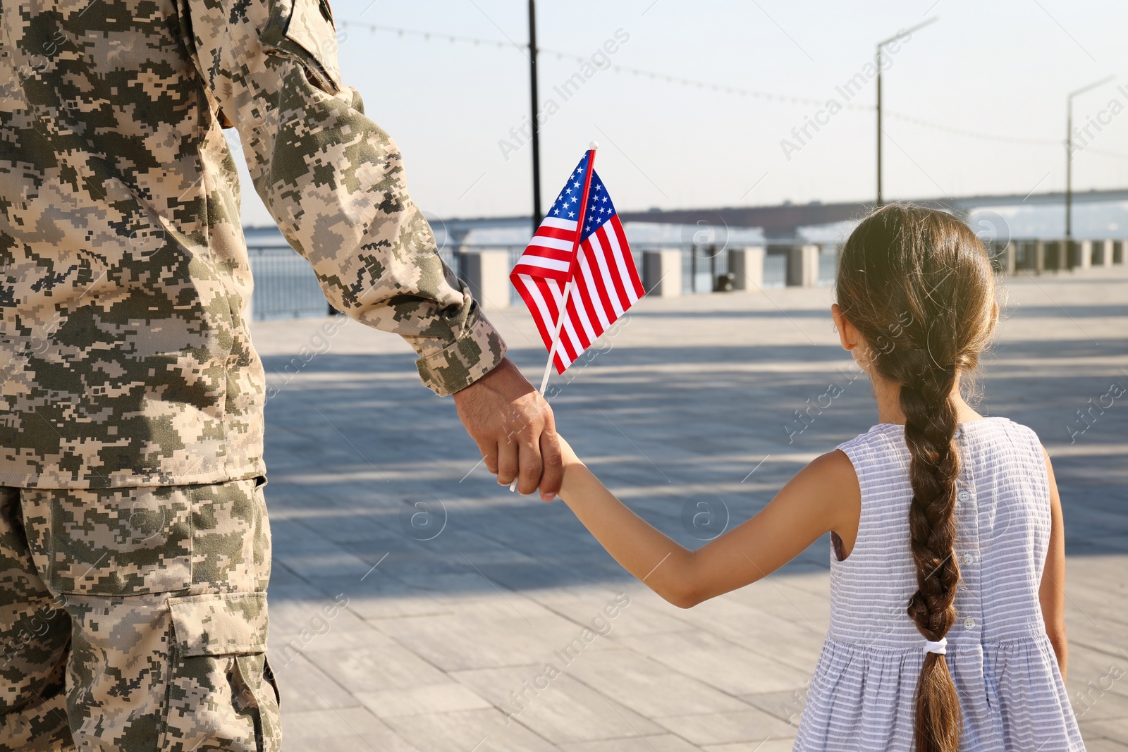 Photo of Soldier and his little daughter with American flag holding hands outdoors, back view. Veterans Day in USA