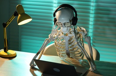 Photo of Waiting concept. Human skeleton sitting at wooden table with laptop and headphones indoors