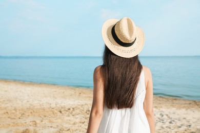 Beautiful young woman wearing straw hat on beach, back view. Stylish headdress