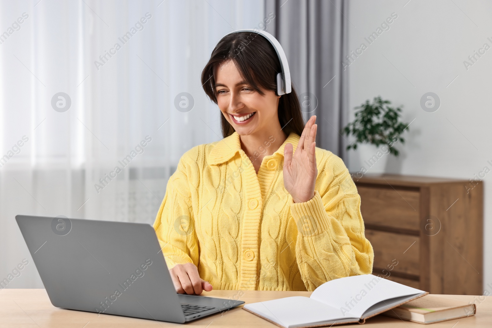 Photo of Happy woman waving hello during video chat via laptop at table indoors