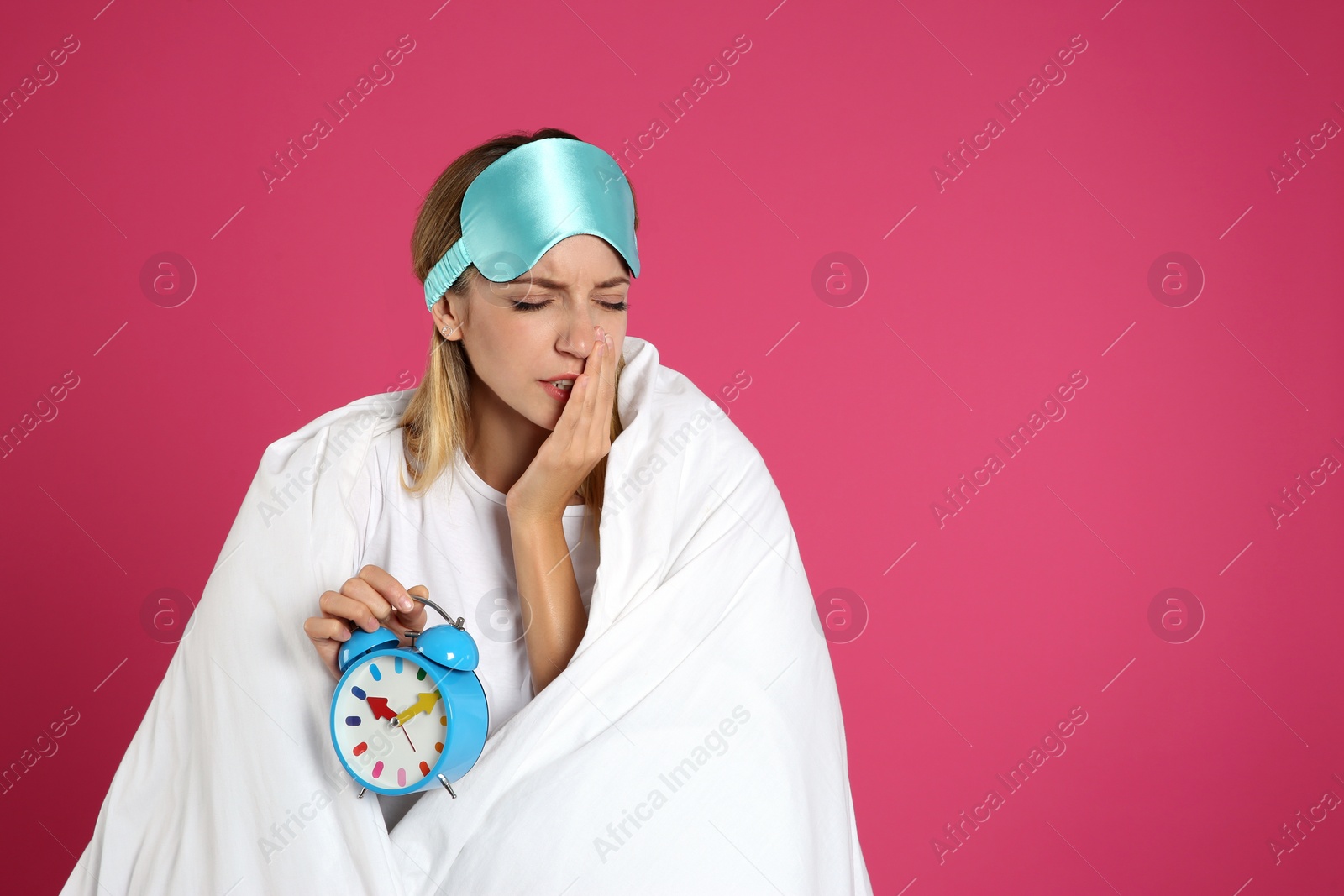 Photo of Young woman in sleeping mask wrapped with blanket holding alarm clock on pink background. Space for text