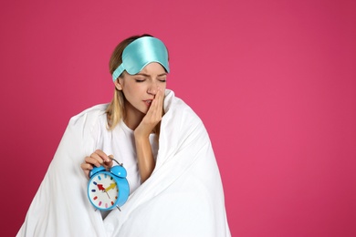 Young woman in sleeping mask wrapped with blanket holding alarm clock on pink background. Space for text