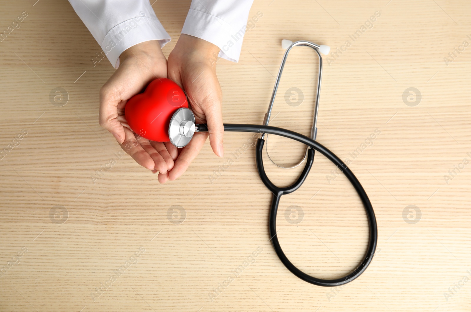 Photo of Doctor holding red heart and stethoscope on wooden background, top view. Cardiology concept