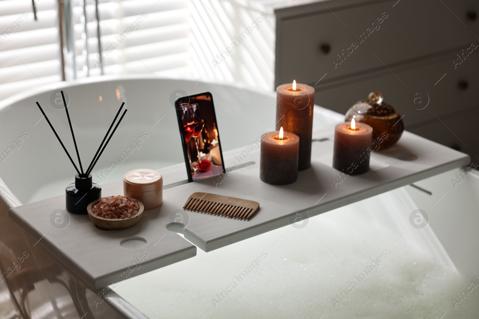 Photo of White wooden tray with smartphone, burning candles and beauty products on bathtub in bathroom