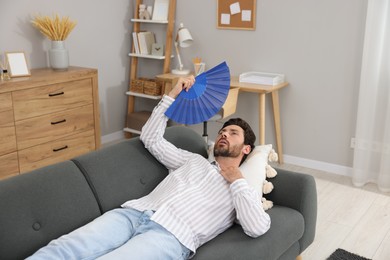 Bearded man waving blue hand fan to cool himself on sofa at home