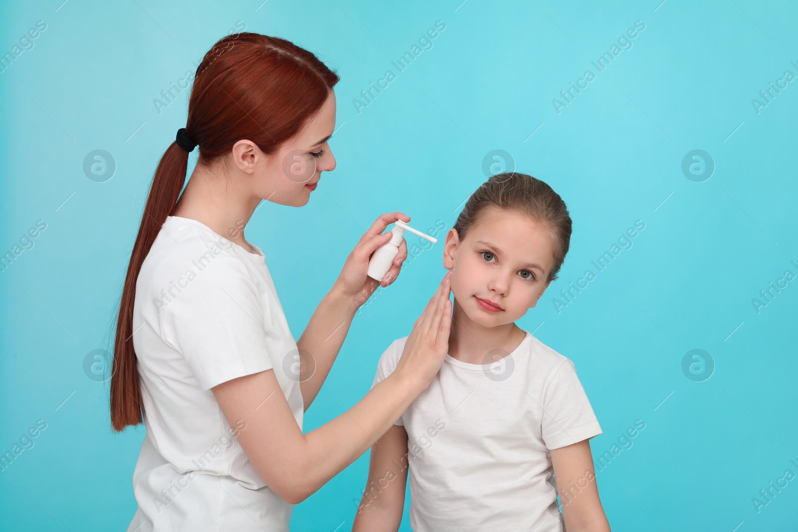 Photo of Mother spraying medication into daughter's ear on light blue background