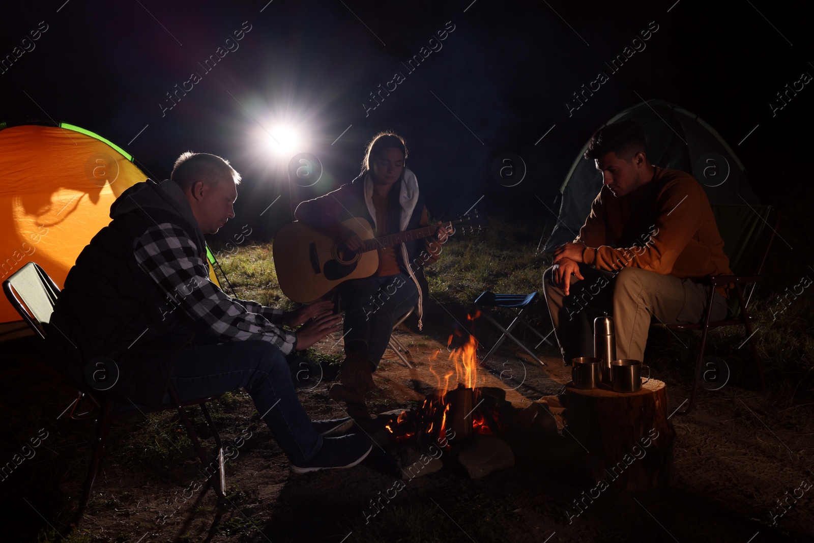 Photo of Group of friends with guitar near bonfire and camping tent outdoors at night