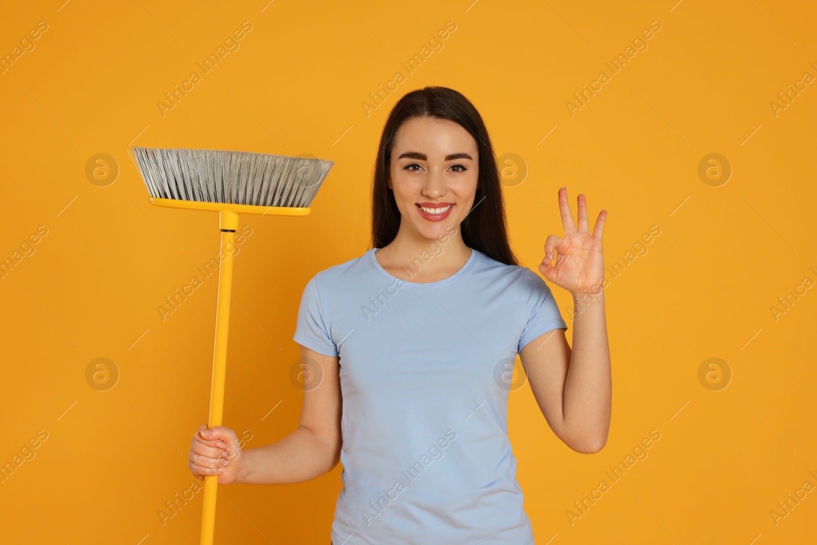 Photo of Beautiful young woman with broom showing OK gesture on yellow background