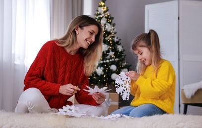 Photo of Happy mother and daughter making paper snowflakes near Christmas tree at home