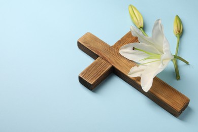 Photo of Wooden cross and lily flowers on light blue background, space for text. Easter attributes