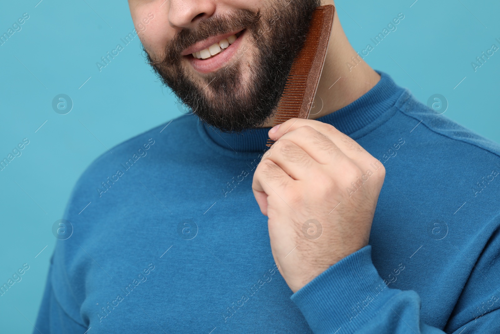 Photo of Handsome young man combing beard on light blue background, closeup