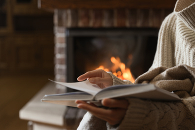 Woman reading book near burning fireplace at home, closeup