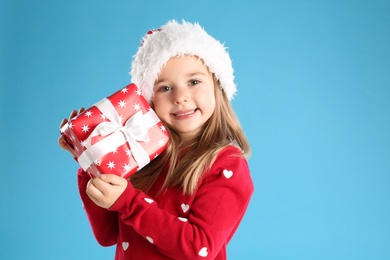 Photo of Cute child in Santa hat with Christmas gift on light blue background