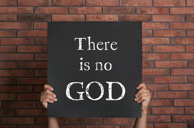 Image of Atheism. Man holding sign with text There Is No God near brick wall, closeup