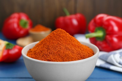 Bowl with aromatic paprika powder and fresh bell peppers on blue table, closeup