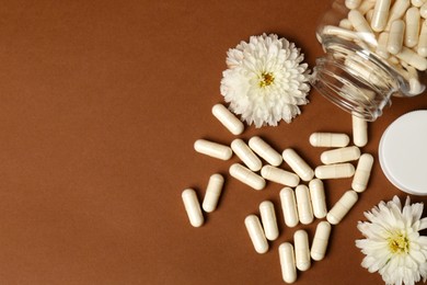 Open medicine bottle, scattered pills and flowers on brown background, flat lay. Space for text