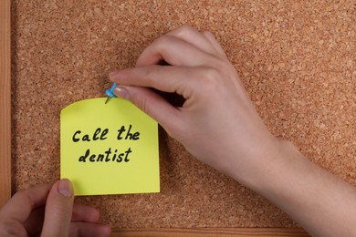 Photo of Woman pinning paper note with phrase Call The Dentist to cork board, closeup