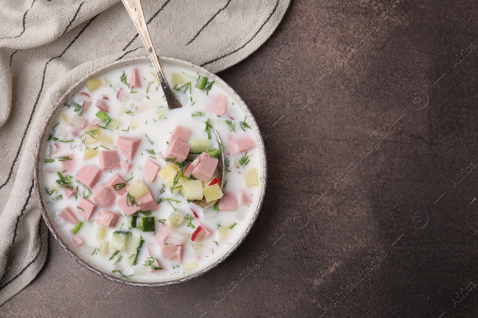 Photo of Delicious cold summer soup (okroshka) with boiled sausage and spoon in bowl on brown table, top view. Space for text