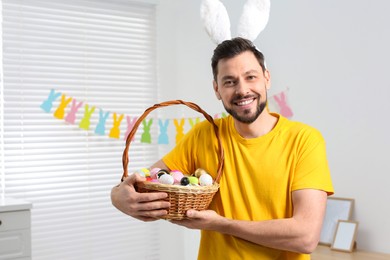 Happy man in bunny ears headband holding wicker basket with painted Easter eggs indoors