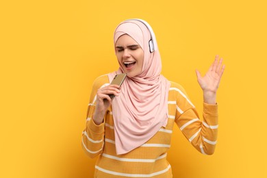 Muslim woman in hijab and headphones singing with smartphone on orange background