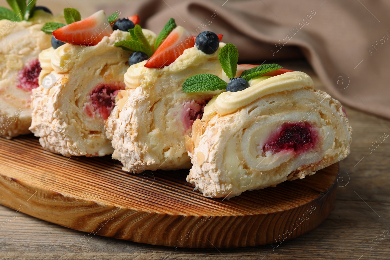 Photo of Pieces of tasty meringue roll with jam, cream, strawberry, blueberry and mint on wooden table, closeup
