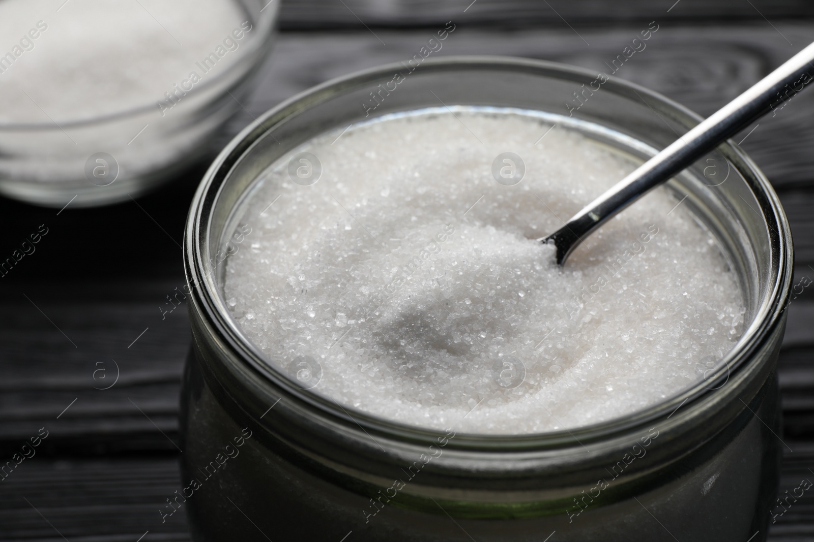 Photo of Granulated sugar and spoon in glass jar on black table, closeup