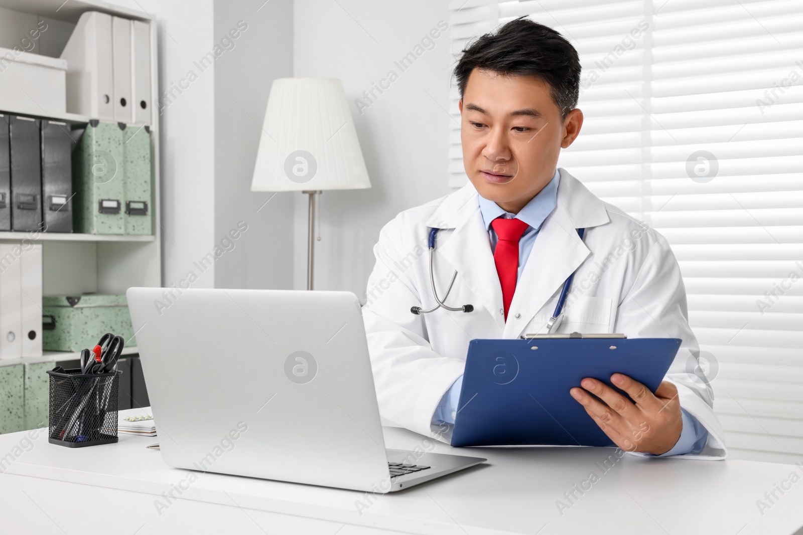Photo of Doctor with laptop and clipboard consulting patient at white desk in clinic. Online medicine