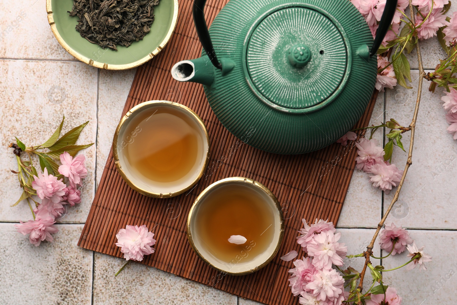 Photo of Traditional ceremony. Cup of brewed tea, teapot, dried leaves and sakura flowers on tiled table, flat lay