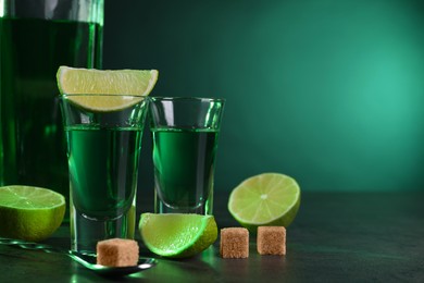 Photo of Absinthe in shot glasses, spoon, brown sugar cubes and lime on gray textured table against green background, space for text. Alcoholic drink