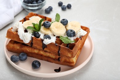 Photo of Plate of delicious Belgian waffles with blueberry, banana, whipped cream and chocolate sauce on light marble table, closeup