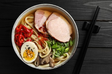 Delicious ramen in bowl and chopsticks on black wooden table, flat lay. Noodle soup