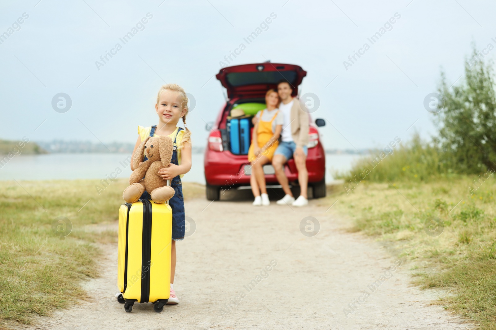 Photo of Little girl with suitcase and toy bunny near car on riverside. Space for text