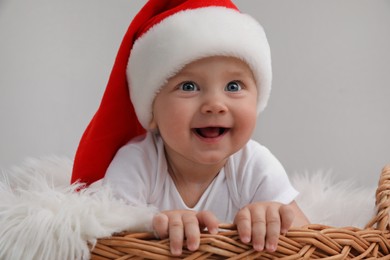 Photo of Cute baby wearing Santa hat in wicker basket on light grey background. Christmas celebration