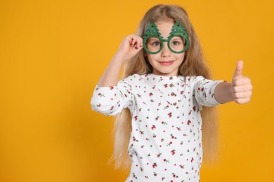 Girl wearing decorative eyeglasses in shape of Christmas trees showing thumb up on orange background. Space for text
