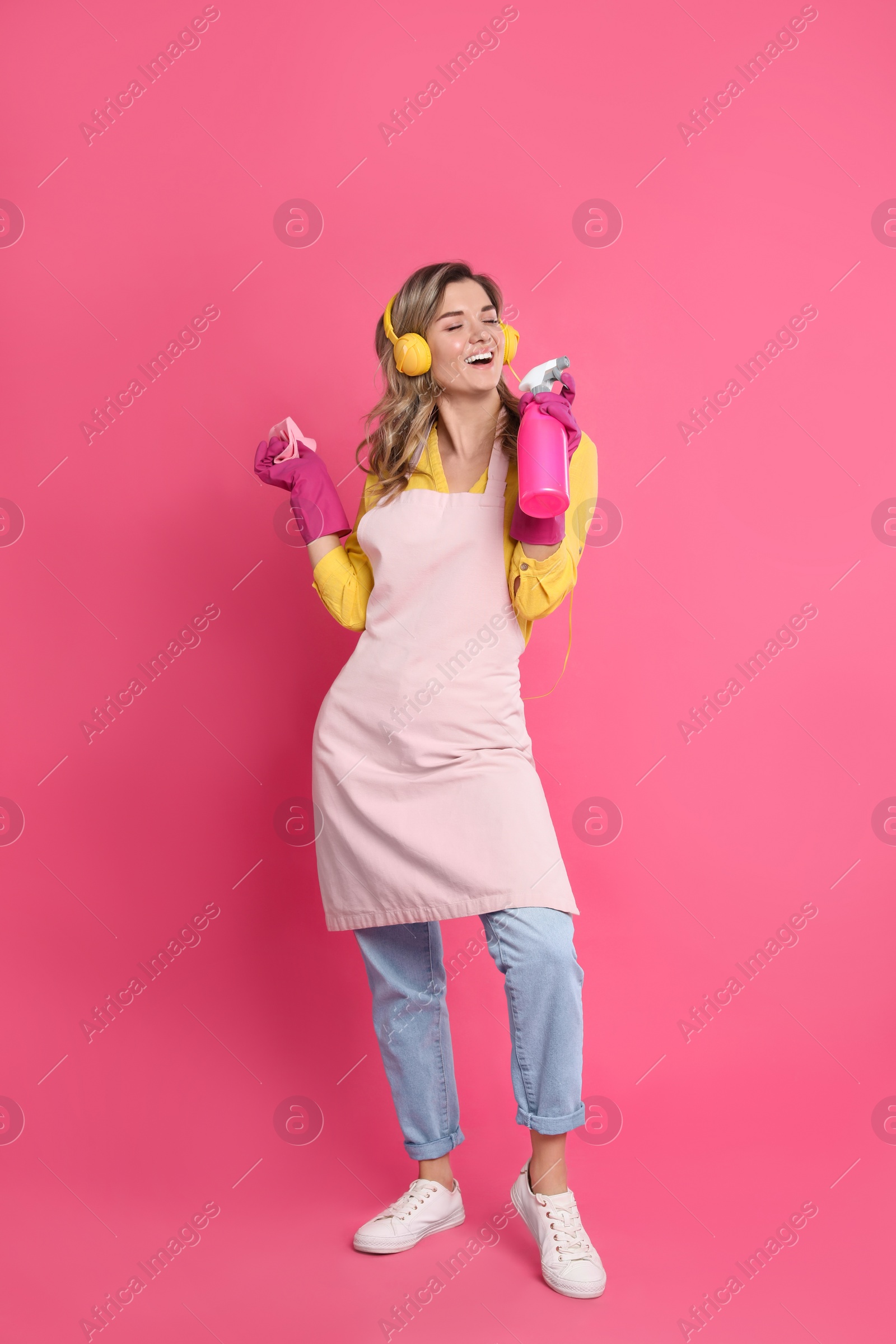 Photo of Beautiful young woman with headphones, rag and bottle of detergent singing on pink background