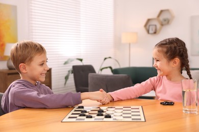 Cute children shaking hands after playing checkers at wooden table in room