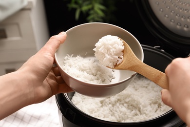 Woman putting tasty rice into bowl from cooker in kitchen, closeup