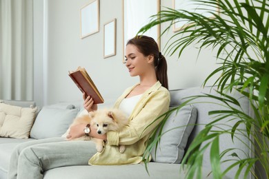 Happy young woman reading book with cute dog on sofa in living room