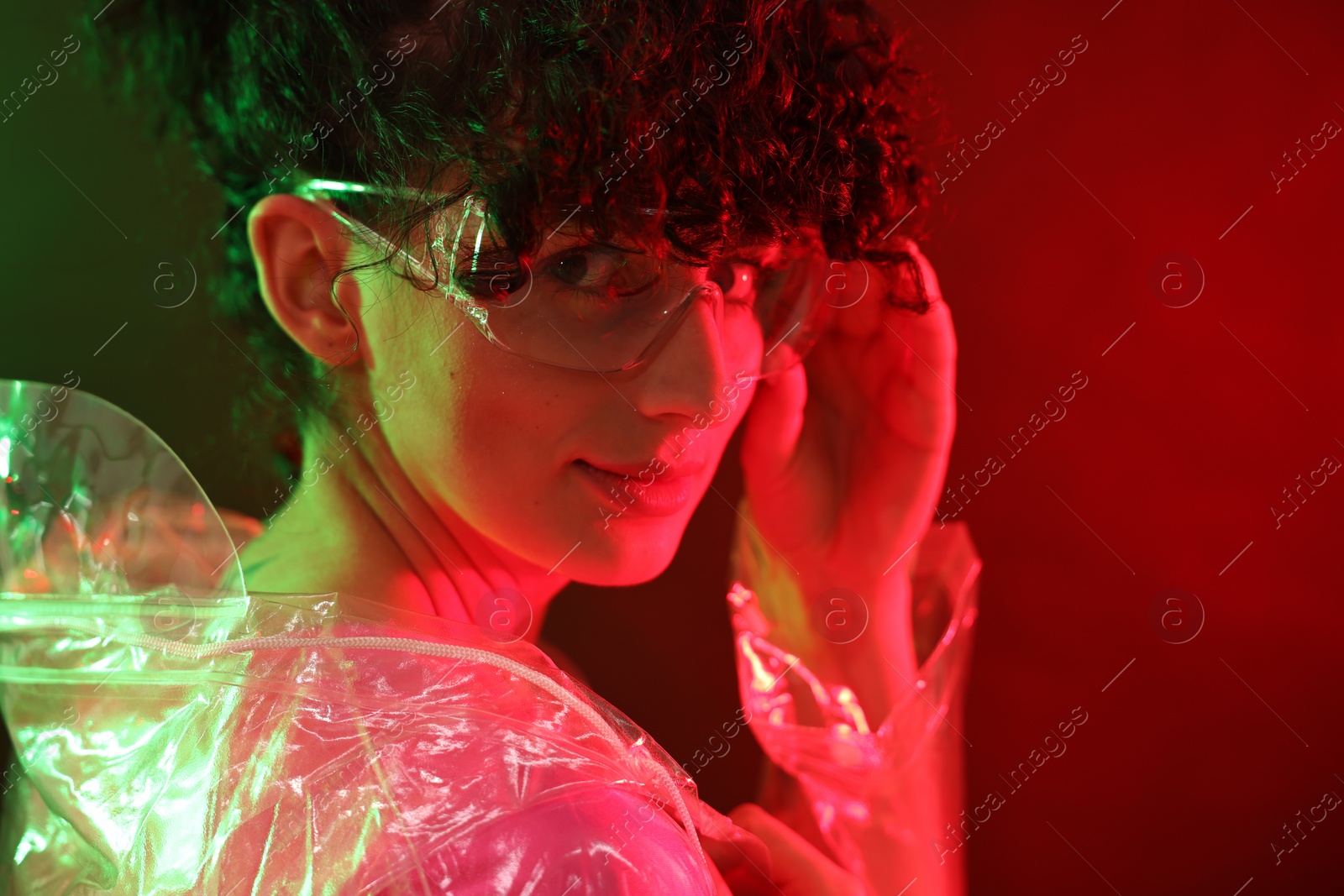 Photo of Beautiful young woman in transparent coat and sunglasses posing on color background in neon lights