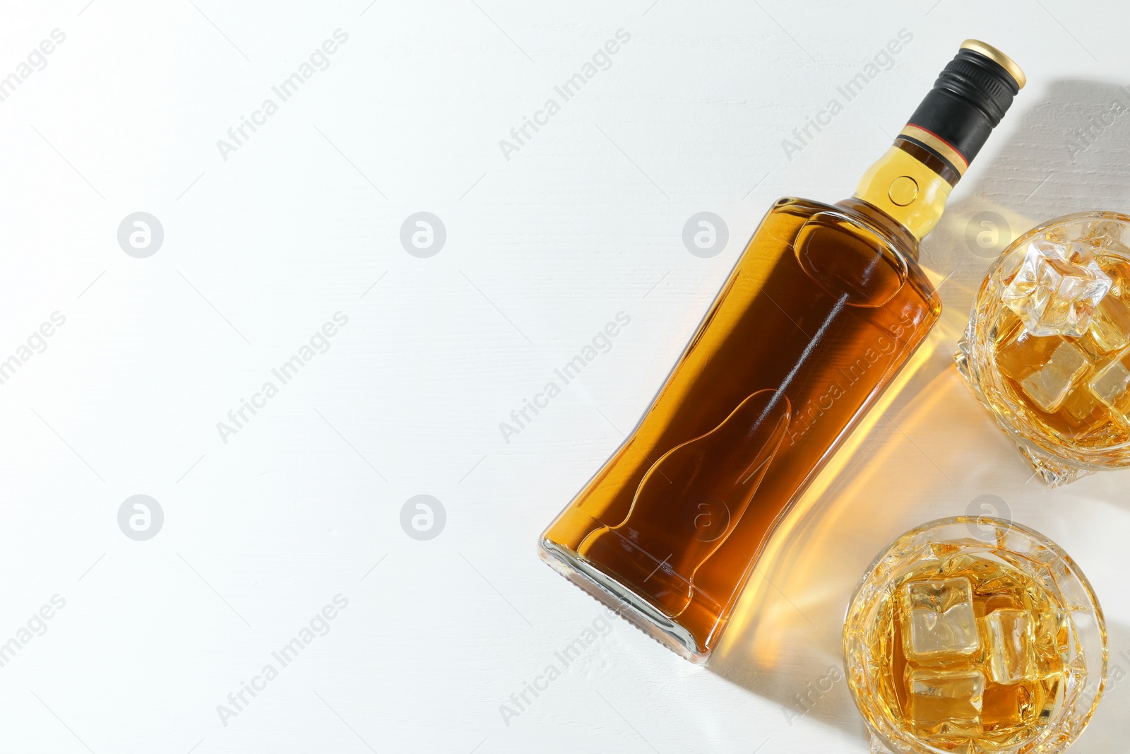 Photo of Whiskey with ice cubes in glasses and bottle on white table, flat lay. Space for text