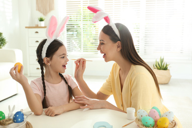 Photo of Happy mother and daughter with bunny ears headbands having fun while painting Easter egg at home