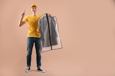 Photo of Dry-cleaning delivery. Happy courier holding garment cover with clothes and pointing at something on beige background, space for text