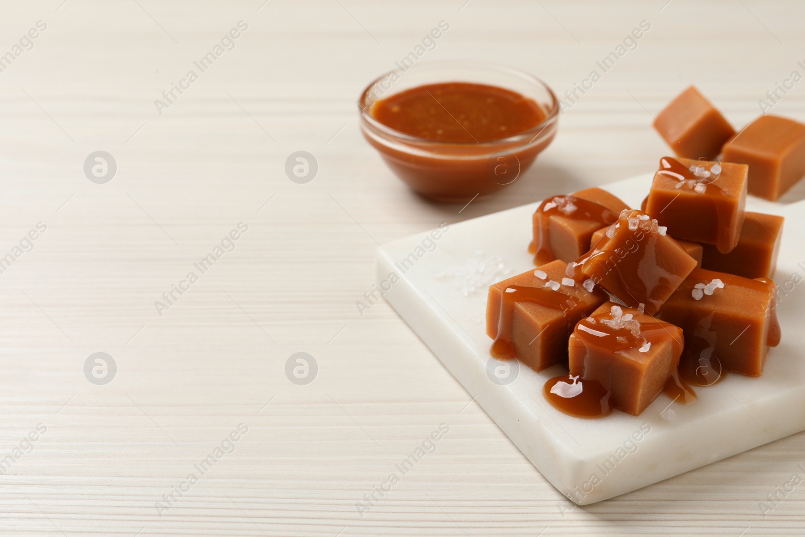 Photo of Tasty caramel candies with sauce and sea salt on white wooden table, space for text