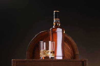Whiskey with ice cubes in glass and bottle on wooden table near barrel against dark background