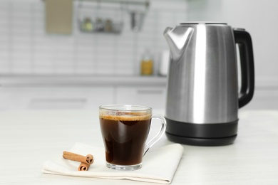 Modern electric kettle, cup of coffee and cinnamon sticks on white wooden table in kitchen