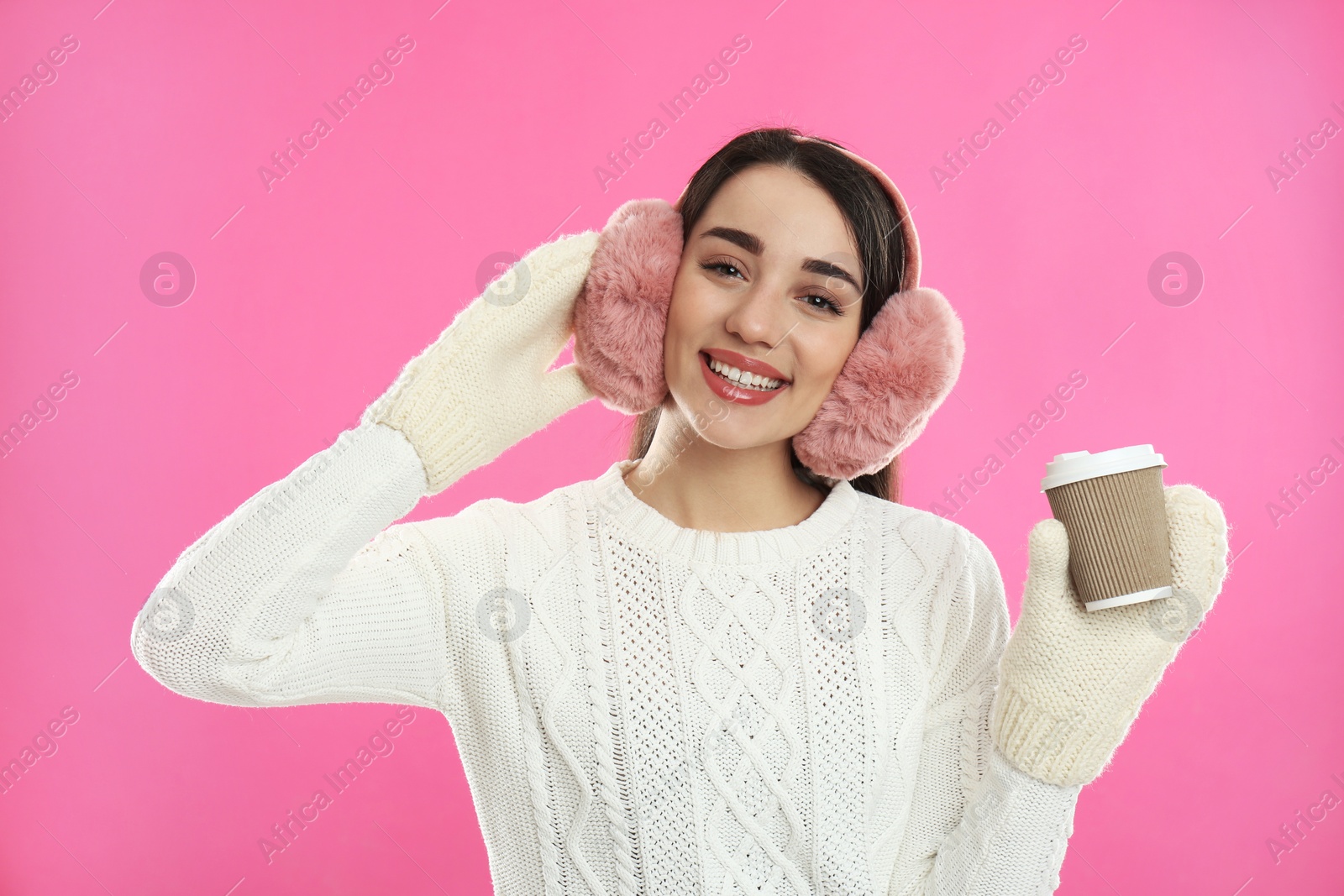 Photo of Beautiful young woman in earmuffs with cup of drink on pink background