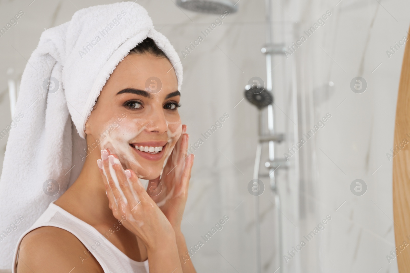 Photo of Happy young woman applying cleansing foam onto face in bathroom
