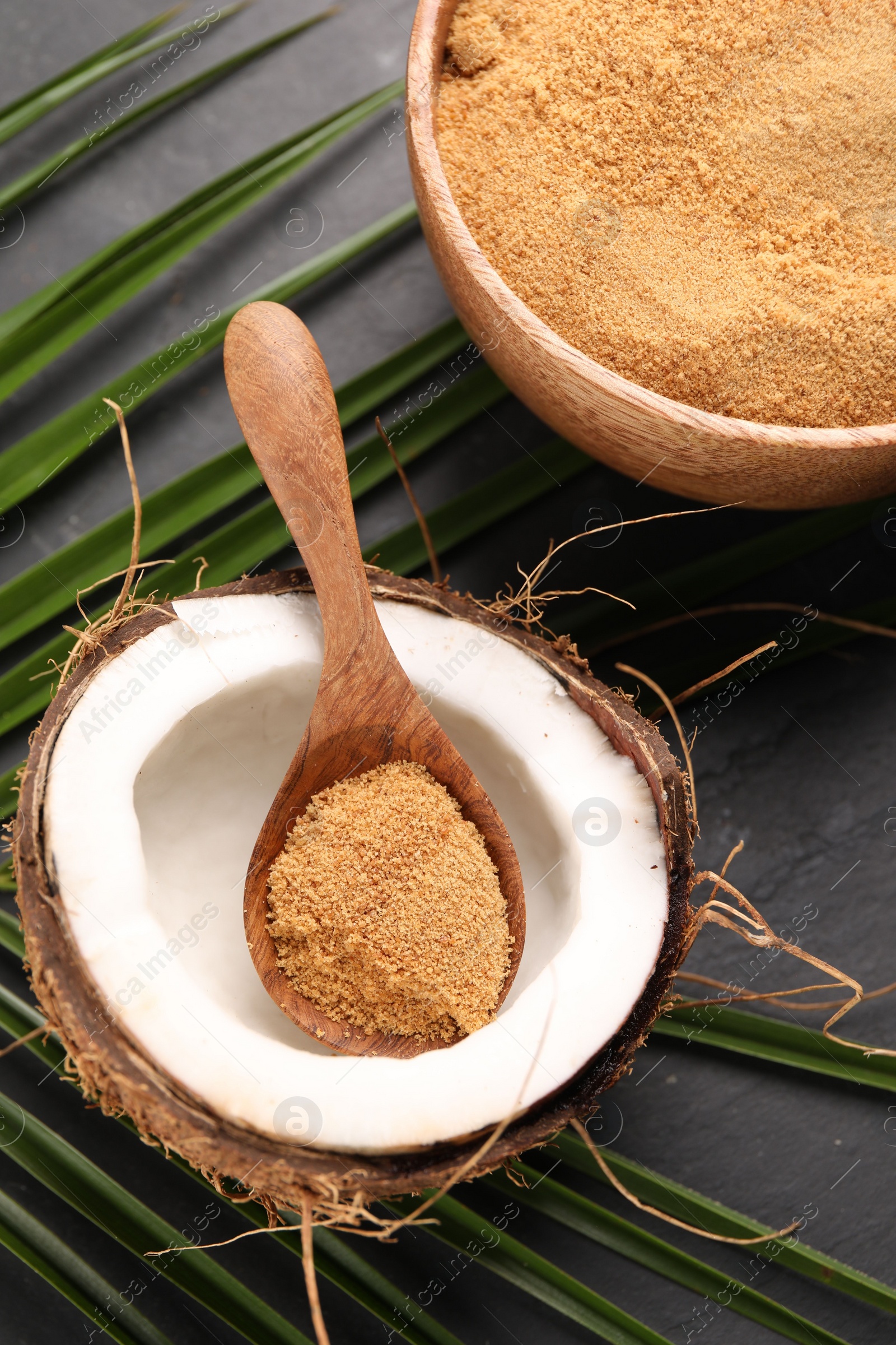 Photo of Spoon with coconut sugar, fruit, bowl and palm leaves on dark textured table, above view