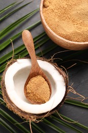 Photo of Spoon with coconut sugar, fruit, bowl and palm leaves on dark textured table, above view